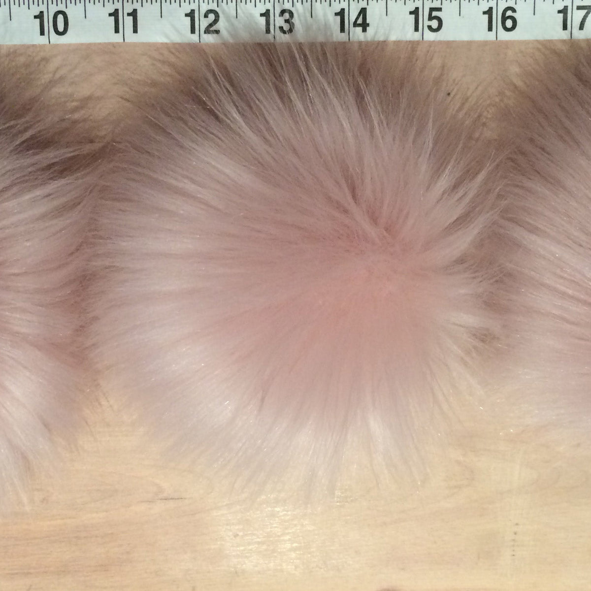 Three fluffy faux fur pom-poms in light pink and white shades, laid out side-by-side on a light wood surface with a measuring tape visible at the top.