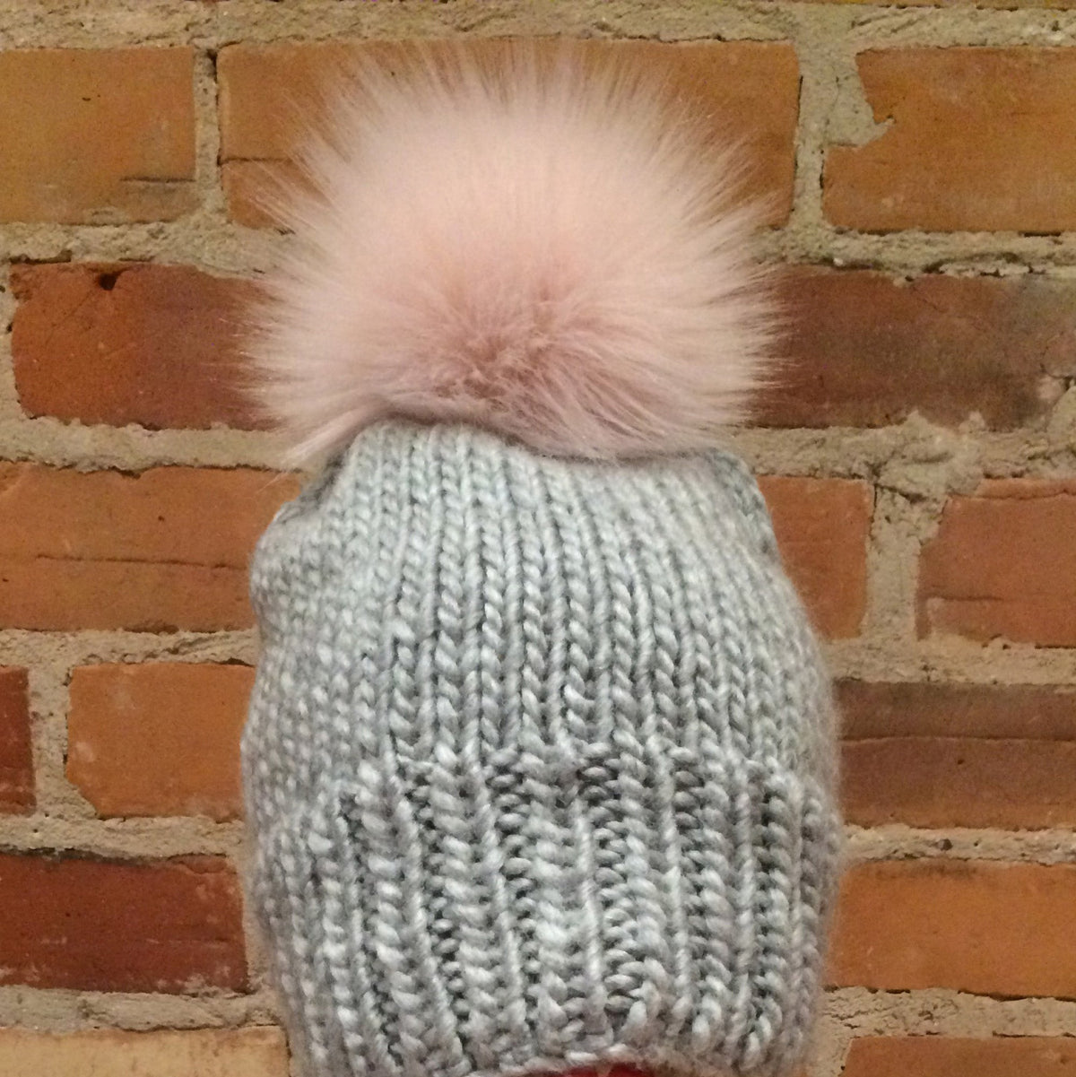 A light blue-gray chunky knit beanie with a fluffy pale pink pom-pom, displayed on a mannequin head against a red-brown brick wall background.