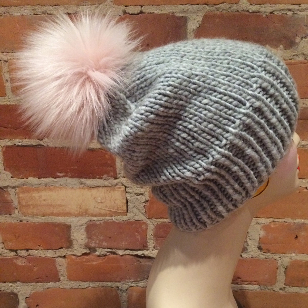 A light gray ribbed knit beanie with a large fluffy pale pink pom-pom, displayed on a mannequin head against a red brick wall background.