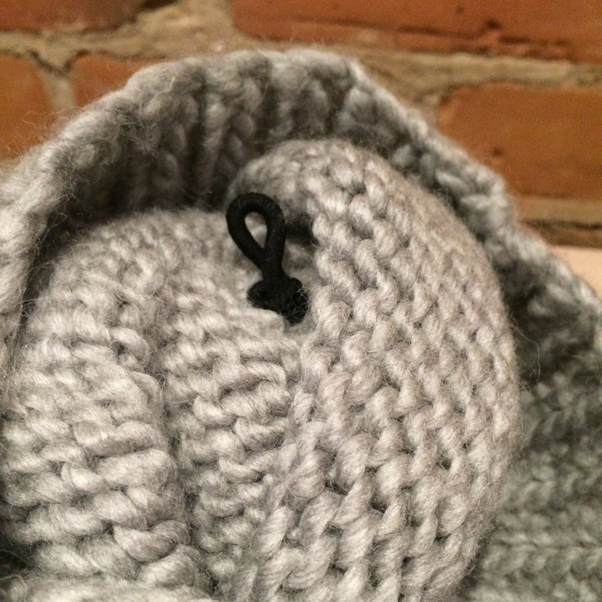 Close-up of a chunky knit gray hat with a black plastic hook fastener, set against a blurred brick background.