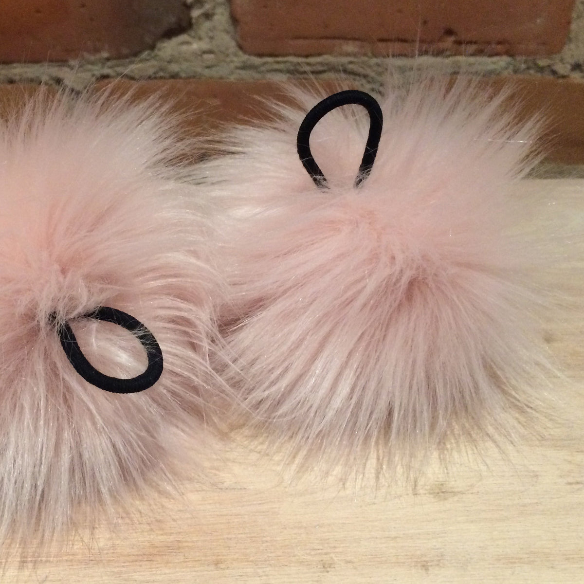 Two fluffy blush pink faux-fur hair accessories with black elastic loops, resting on a light wood surface against a brick wall background.