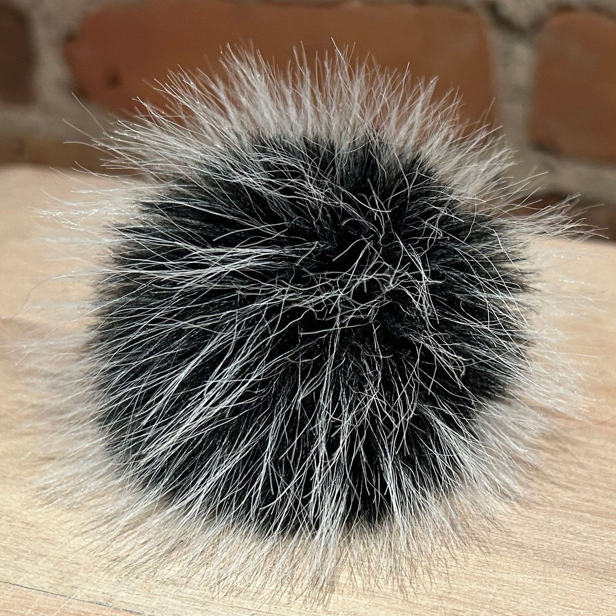 A round black and white faux fur pom-pom accessory resting on a light wood surface with a blurred brick wall background.