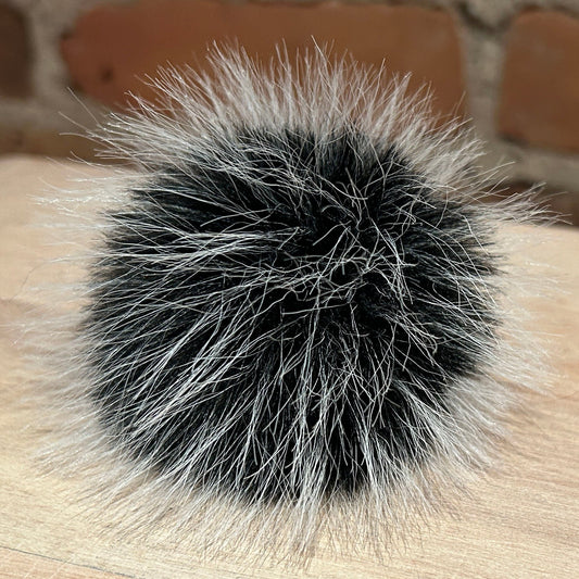 A round black and white faux fur pom-pom accessory resting on a light wood surface with a blurred brick wall background.