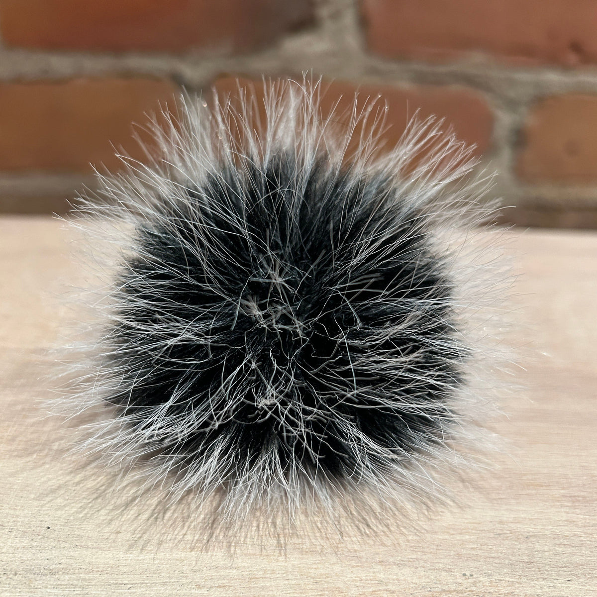 A round fluffy pom-pom accessory made of black and silver-gray faux fur, resting on a light wood surface with a blurred brick wall background.