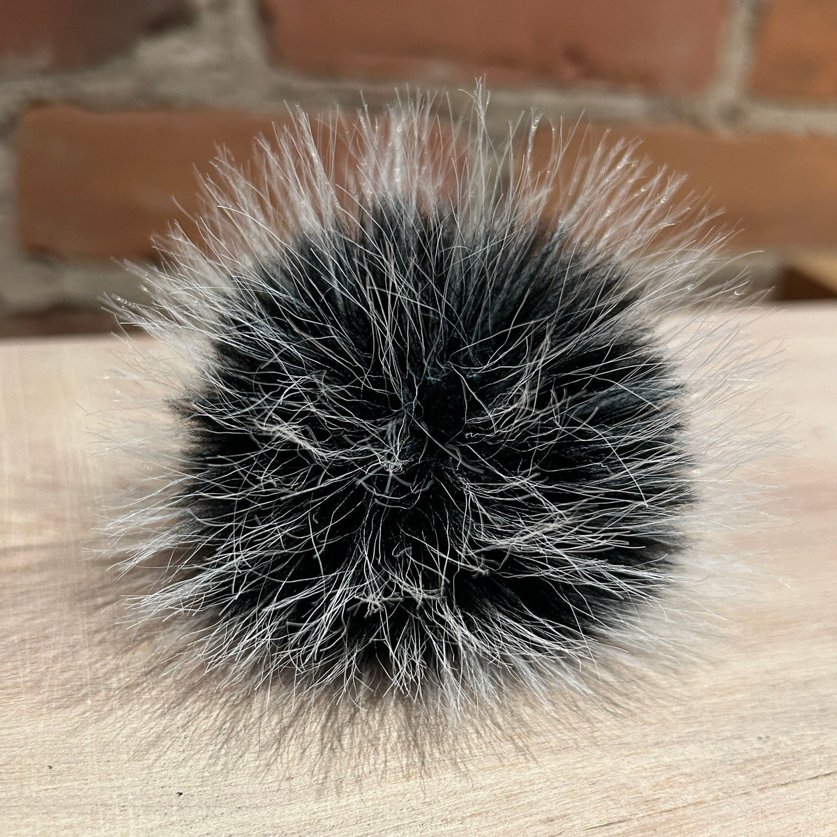 A round fluffy pom-pom accessory made of black and silver-gray faux fur, resting on a light wood surface with a blurred brick wall background.