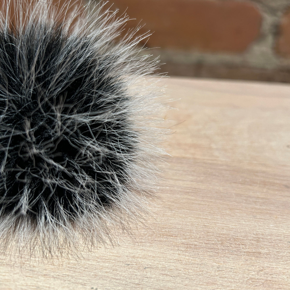A close-up of a fluffy pom-pom accessory made from black and silver-gray faux fur, resting on a light wood surface with a blurred brick wall in the background.