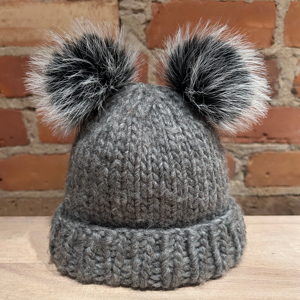 A small gray chunky knit beanie hat with two small black and white faux fur pom-poms on top, displayed on a wooden surface against a brick wall background.