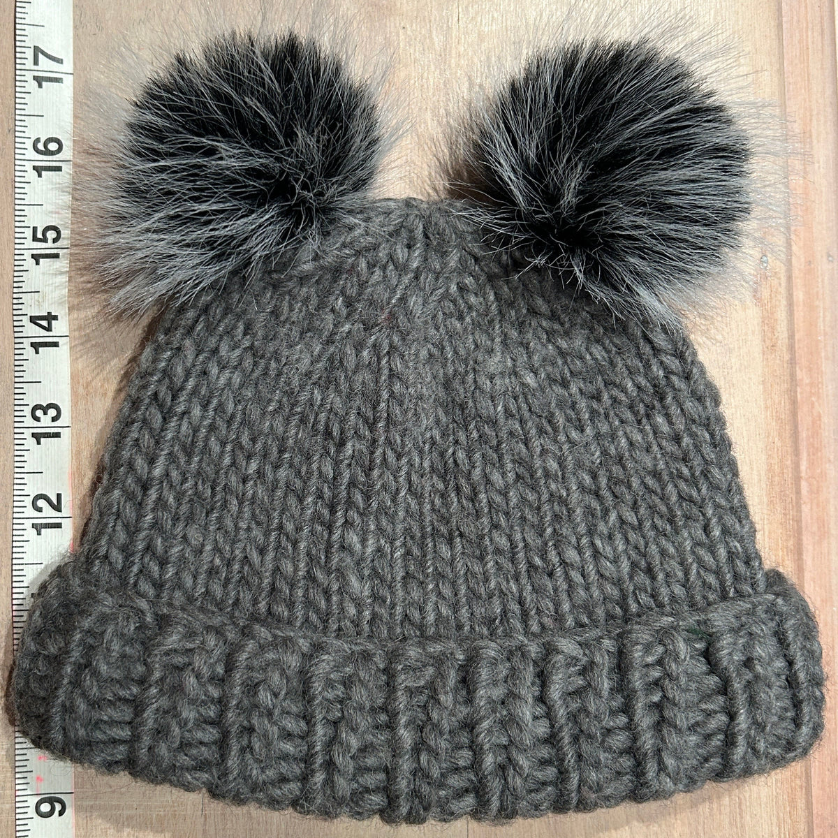 A small gray chunky knit beanie with two small black and gray pom-poms, laid flat on a light wood surface next to a measuring tape.
