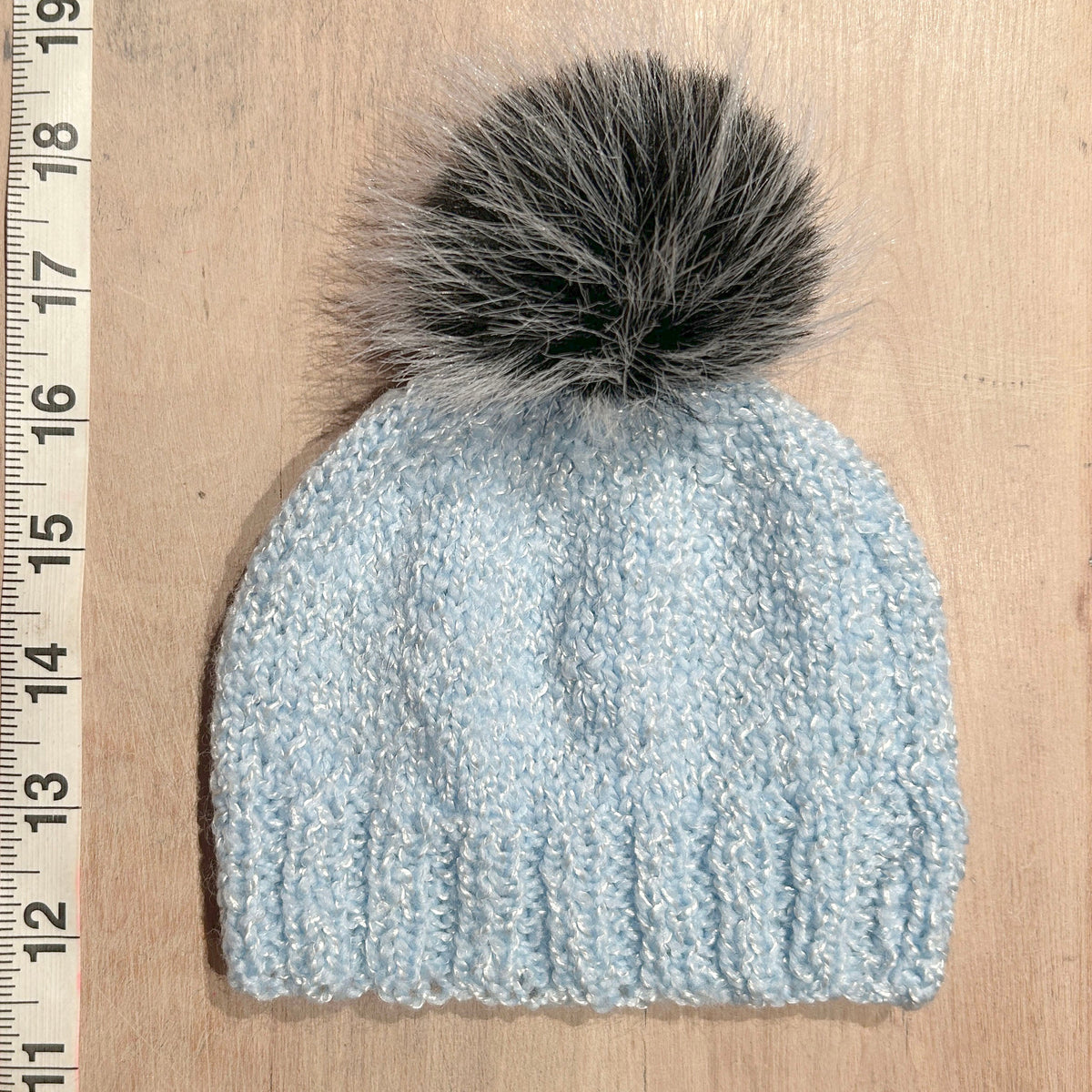 A small light blue knit beanie with a small black and gray faux fur pom-pom, laid flat on a light wood surface next to a wooden ruler for scale.