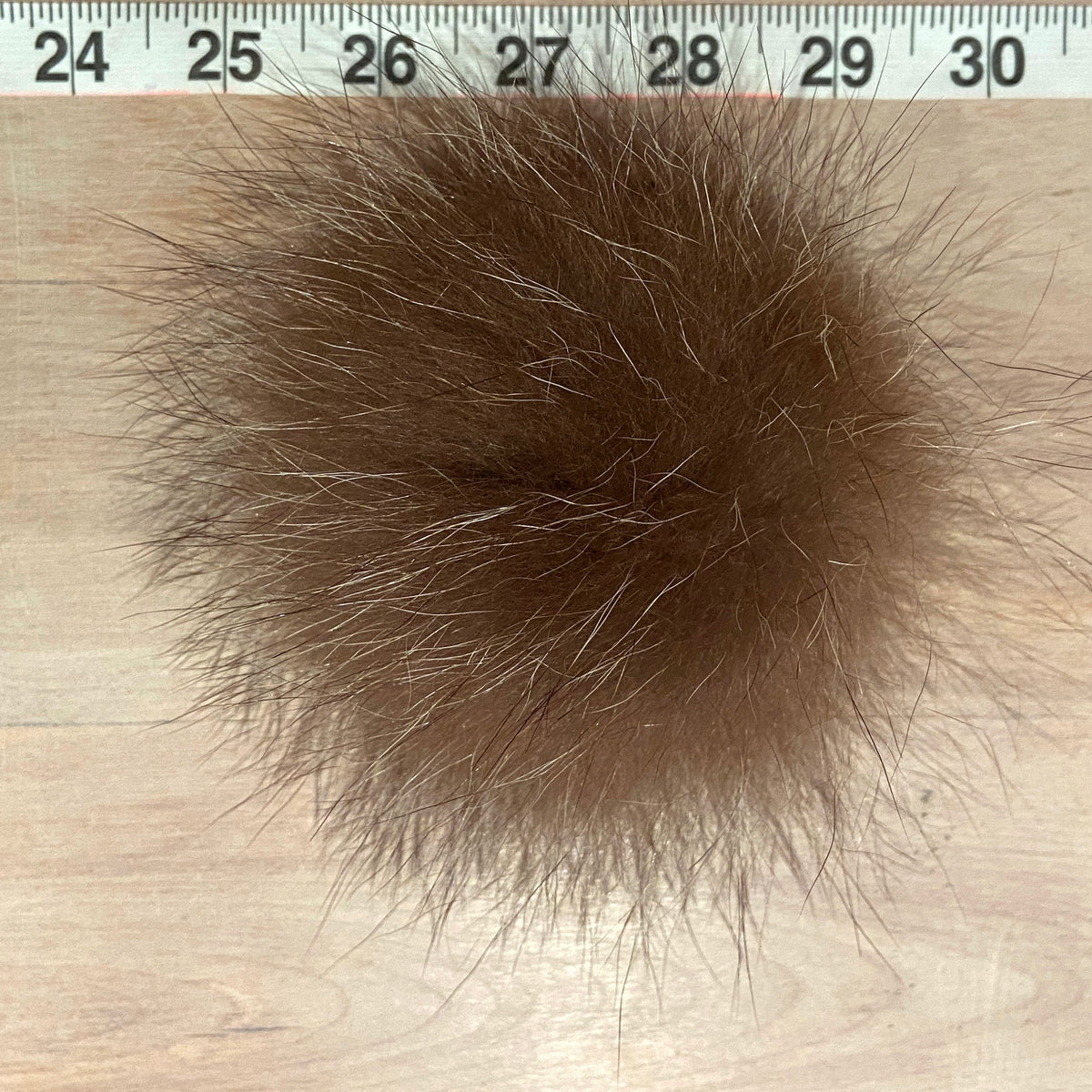 A brown raccoon fur pom-pom ball shown on a light wood surface with a measuring tape ruler at the top for scale.