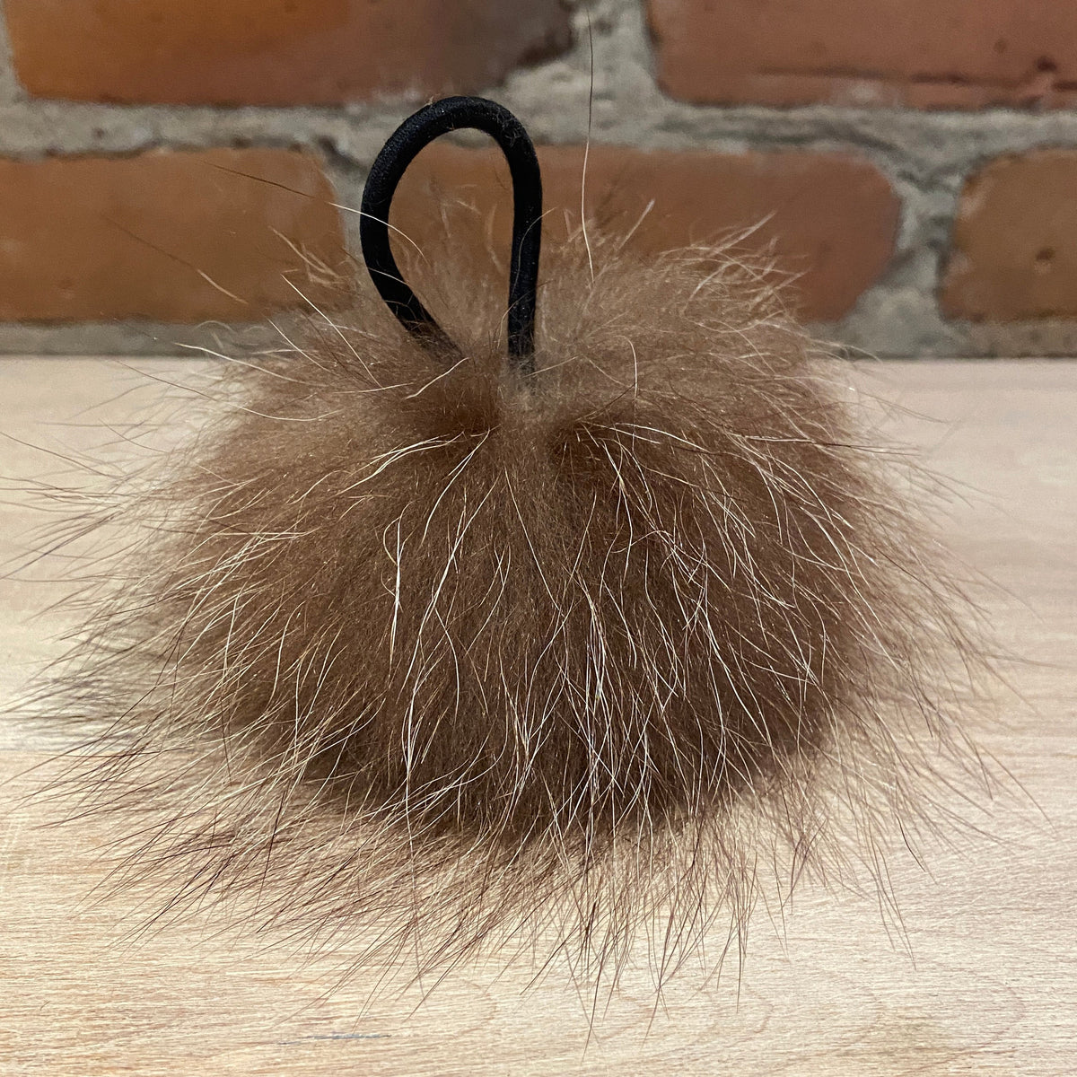 A brown raccoon fur pom-pom keychain with a black elastic loop, resting on a light wood surface with a brick wall background.