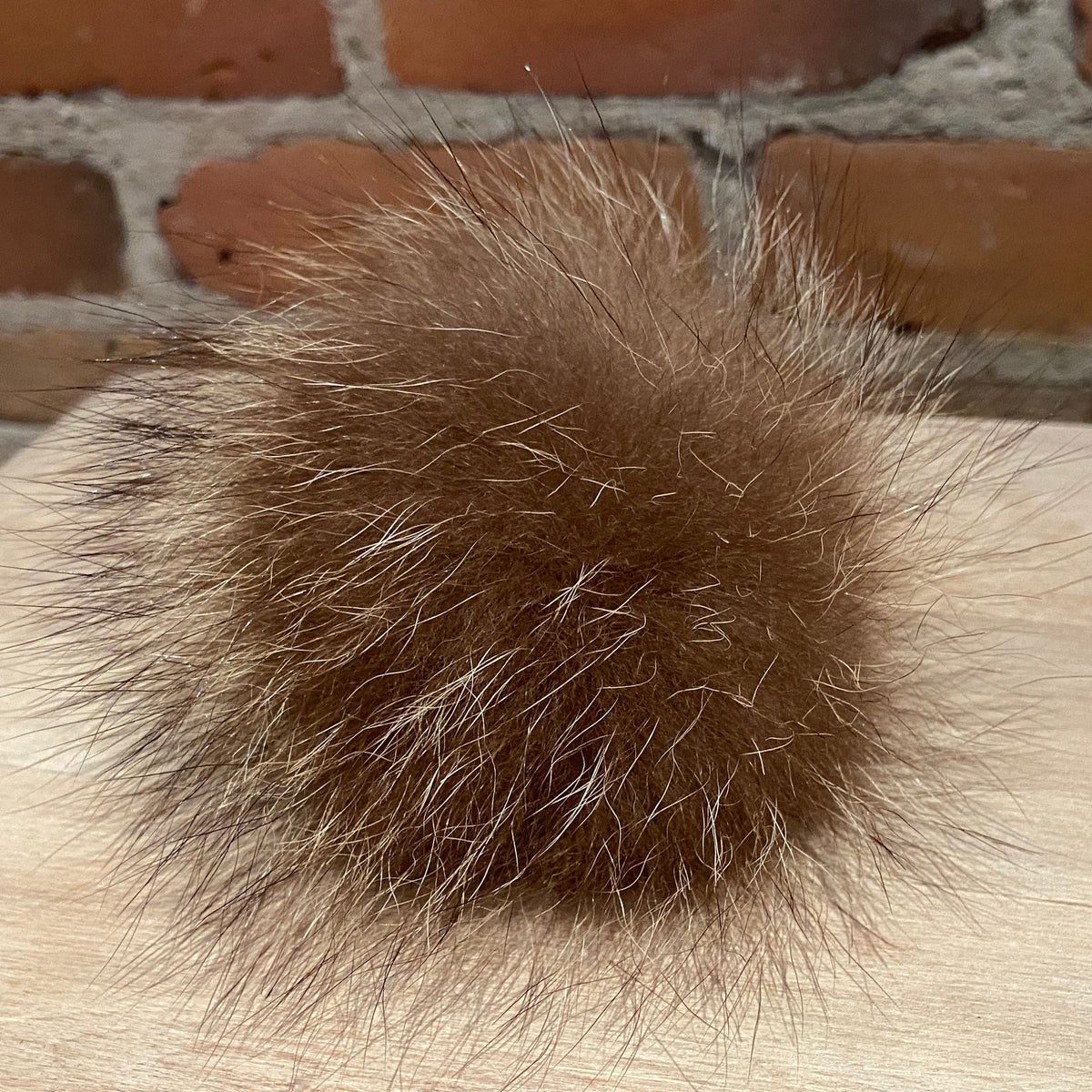 A fluffy brown real fur pom-pom ball resting on a light wood surface with a red brick wall in the background.