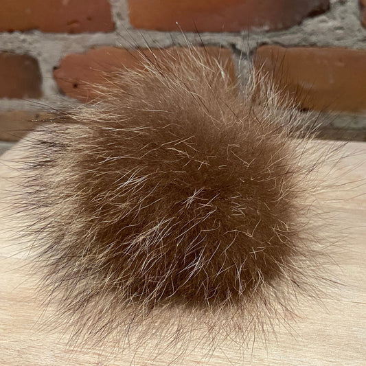 A fluffy brown real fur pom-pom ball resting on a light wood surface with a red brick wall in the background.