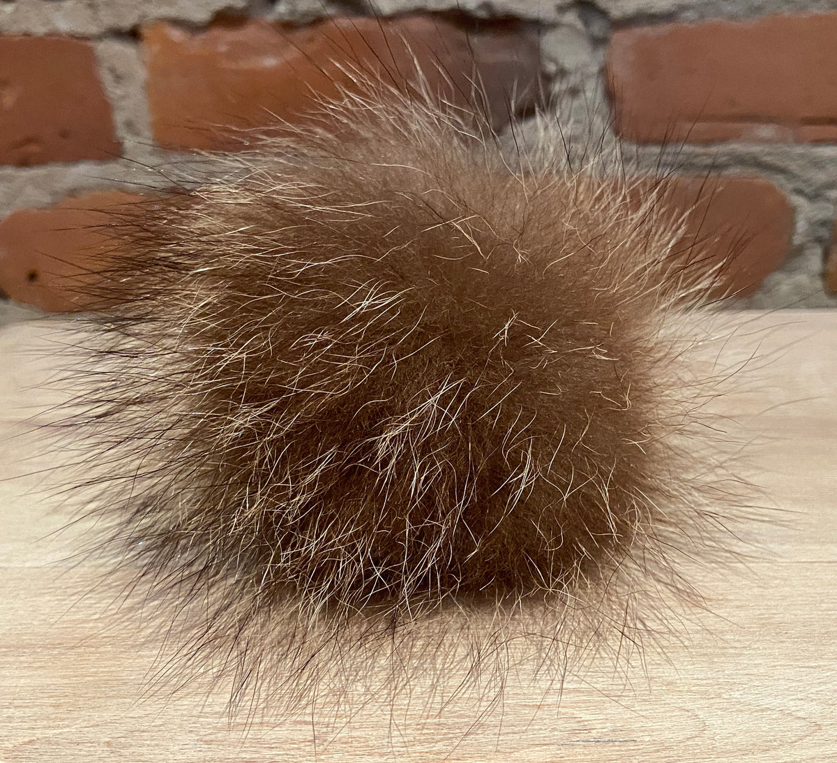 A brown real fur pom-pom ball with natural white and light tan tips, resting on a light wood surface with a blurred brick wall background.