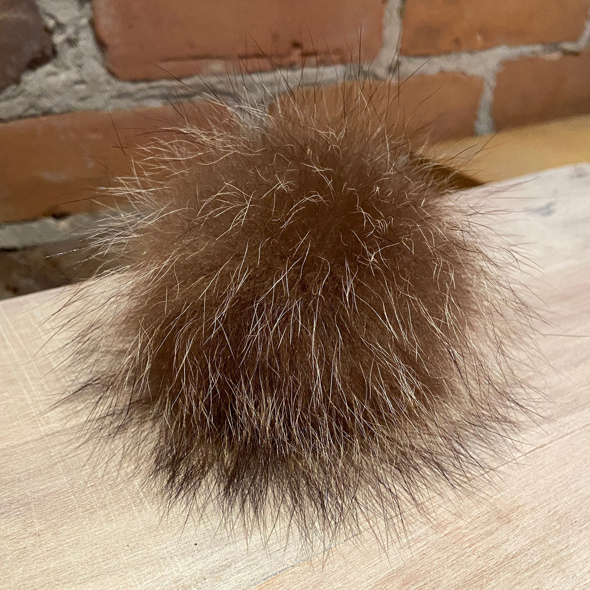 A fluffy brown raccoon fur pom-pom ball resting on a light wood surface with a brick wall in the background.