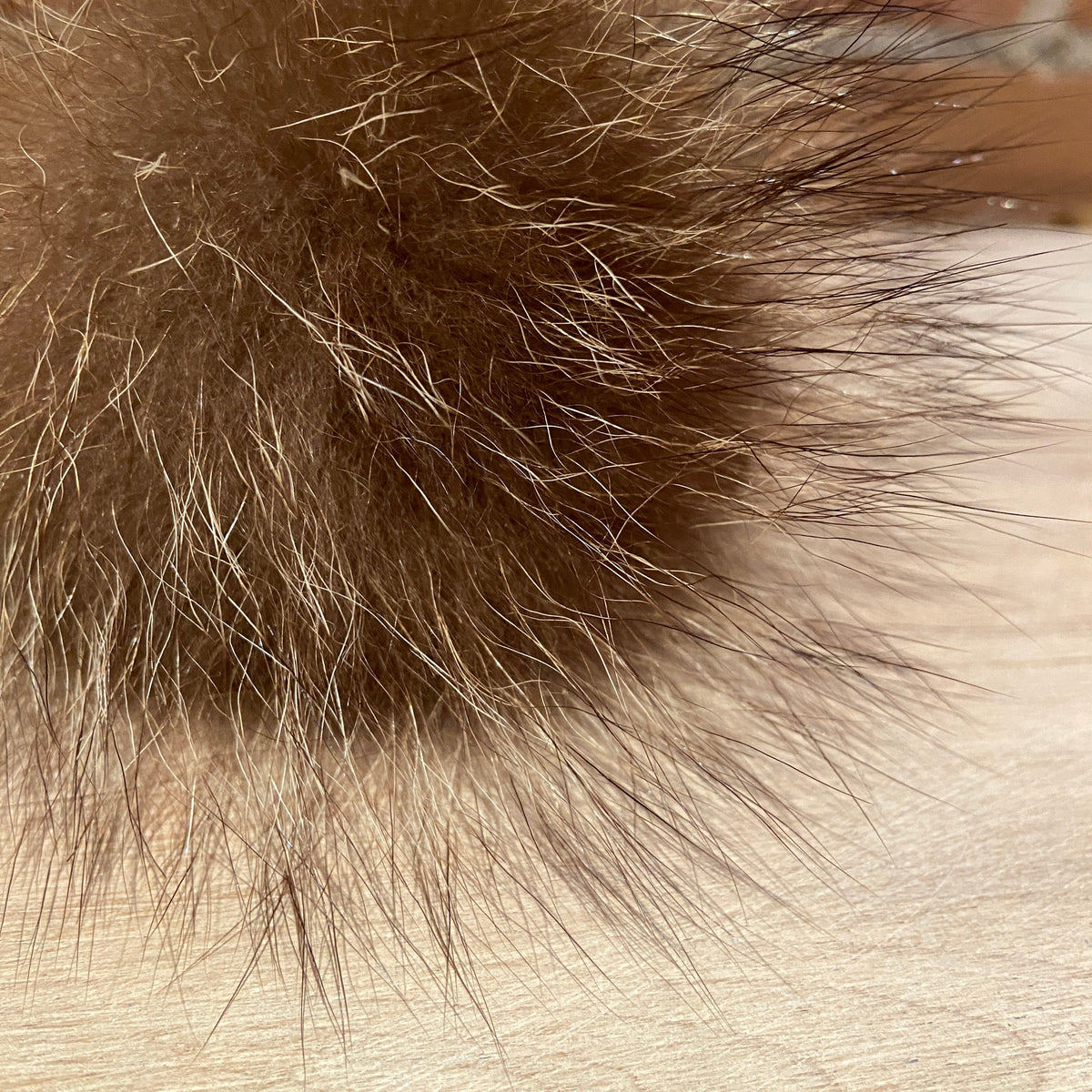 A close-up of a brown real fur pom-pom with mixed dark and light brown fibers, resting on a light wood surface.