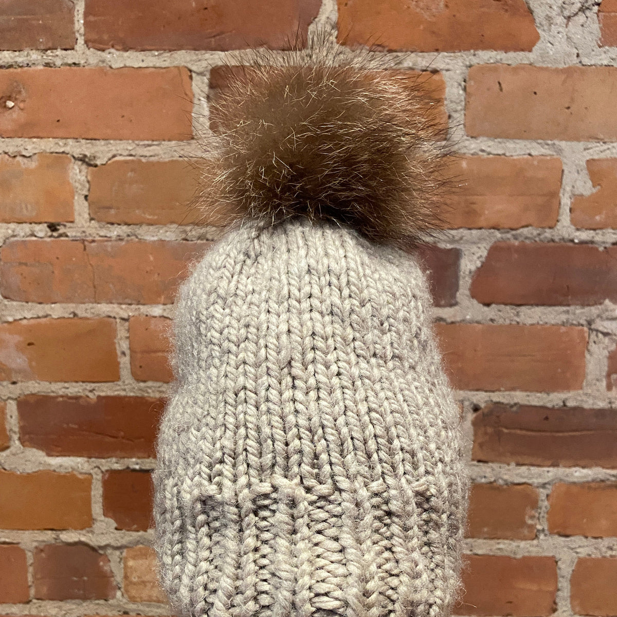 A chunky knit beanie hat in a light oatmeal gray color with a fluffy brown raccoon-fur pom-pom, displayed on a mannequin head against a red brick wall background.