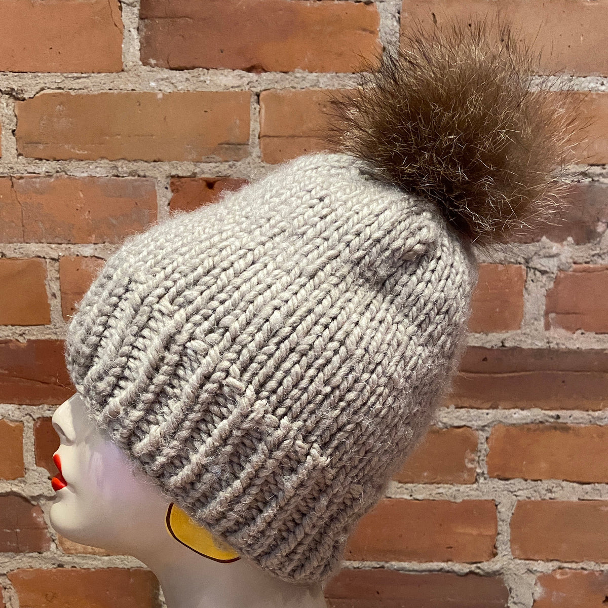 A light gray ribbed knit beanie with a large brown raccoon fur pom-pom, displayed on a mannequin head against a red brick wall background.