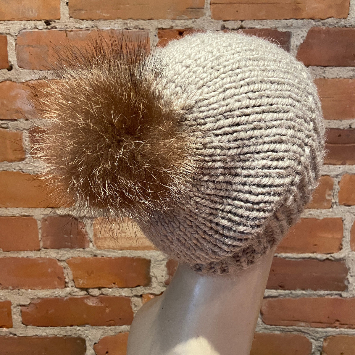 A chunky knit beanie hat in a light oatmeal beige color with a brown real-fur pom-pom, displayed on a mannequin head against a red brick wall background.