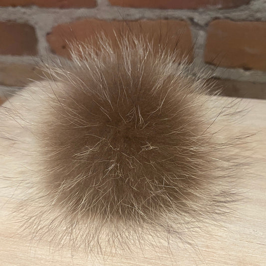A fluffy, round brown raccoon fur pom-pom resting on a light-colored wooden surface with a brick wall background.