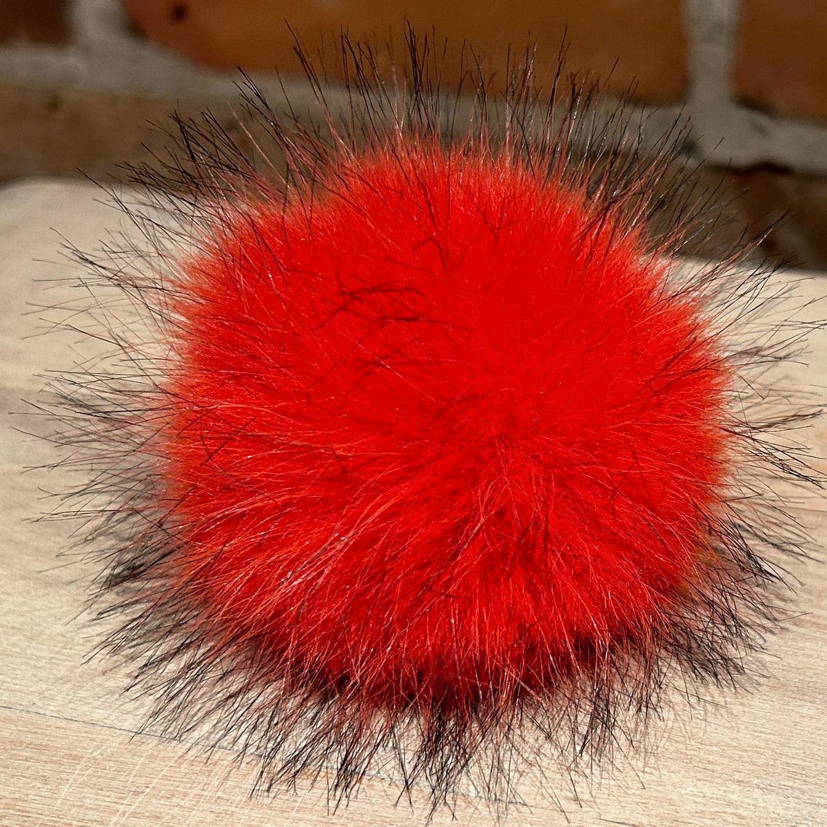 A bright red fluffy pom-pom with dark brown bristles, resting on a light wood surface with a brick wall in the background.