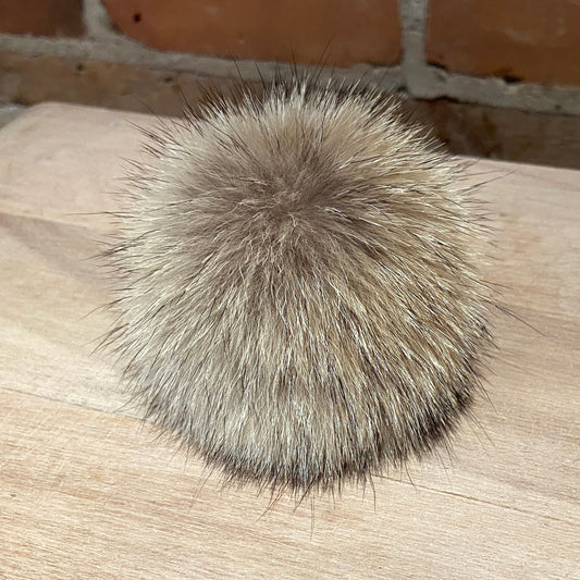 A round, fluffy coyote-fur pom-pom accessory in shades of beige, brown, and gray, resting on a light wood surface with a brick wall background.