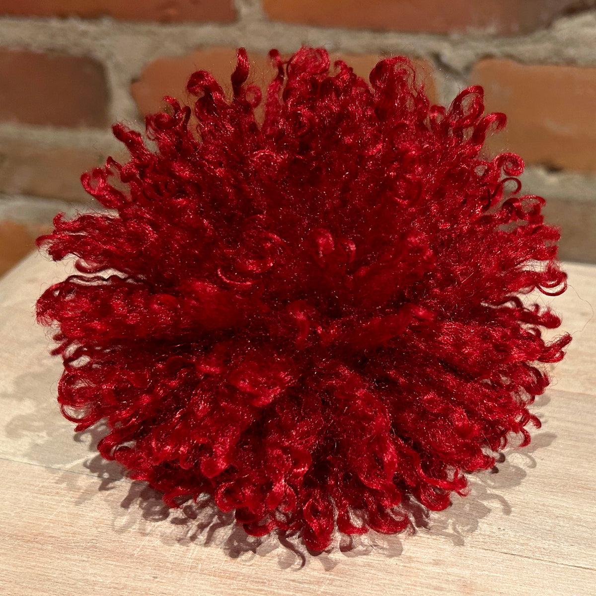 A vibrant deep red curly pom-pom decoration with subtle glitter, resting on a light wood surface with a brick wall background.