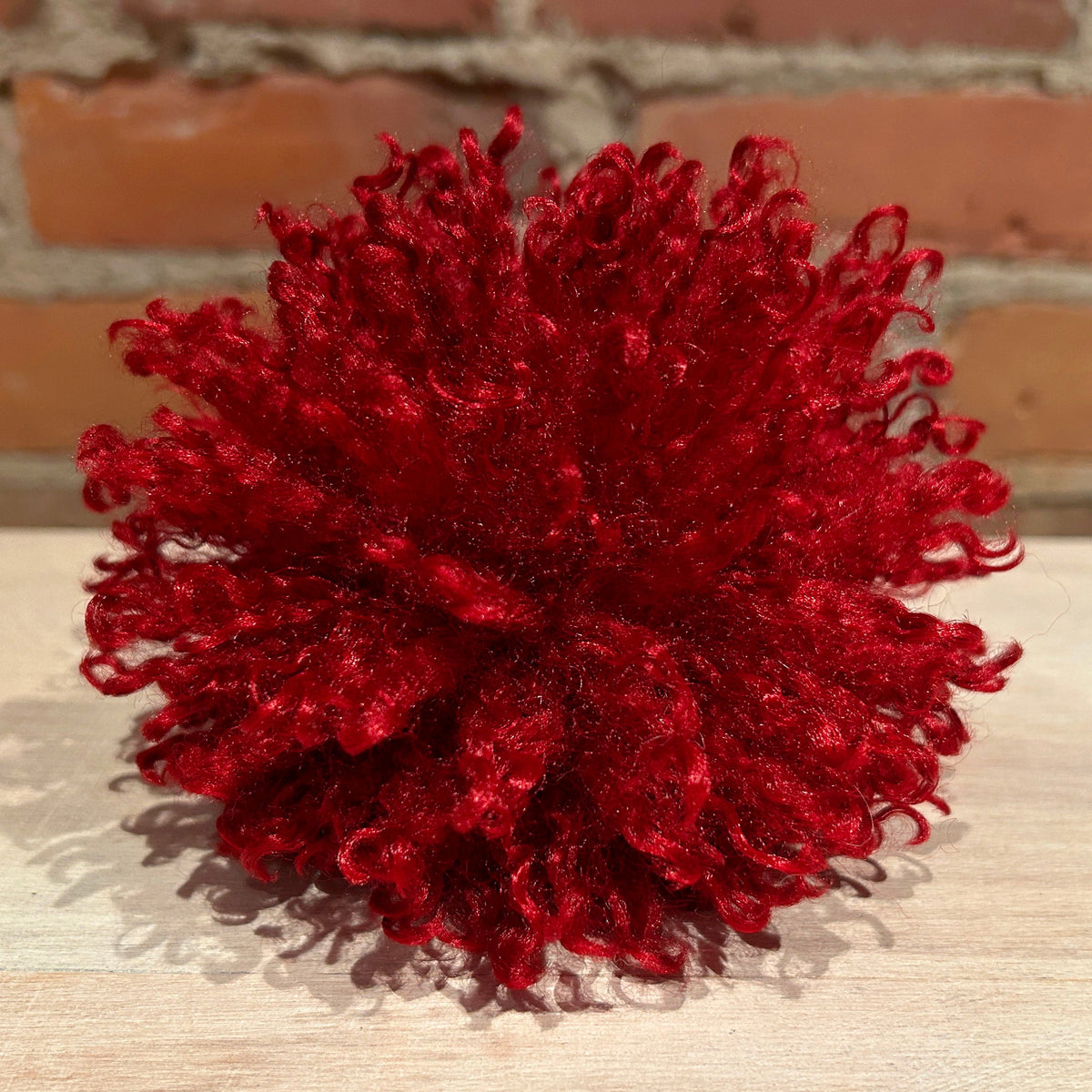 A vibrant deep red curly faux-fur pom-pom decoration, photographed on a light wooden surface with a blurred brick wall background.