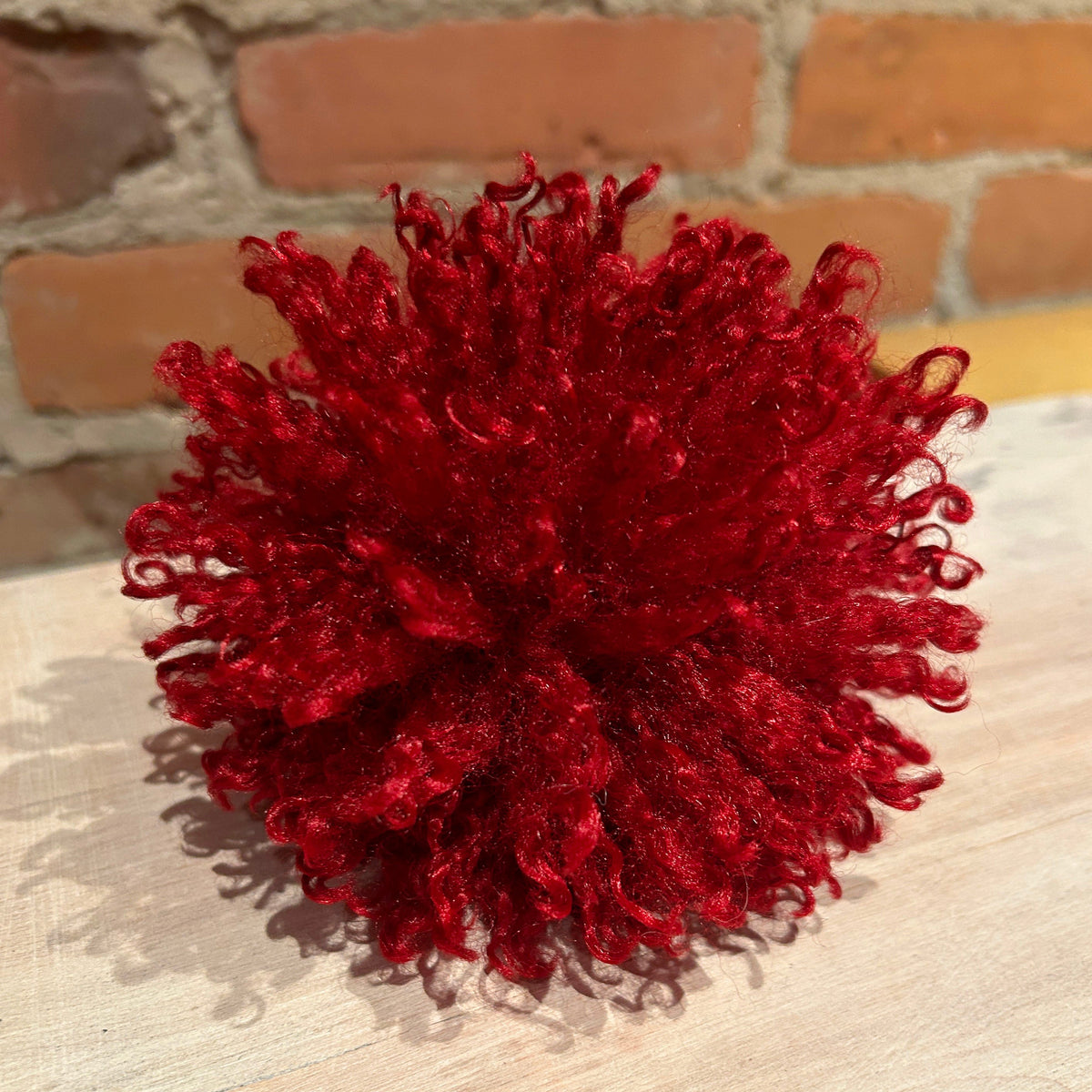A bright red curly pom-pom decoration, resembling curly hair, resting on a light-colored wooden surface with a brick wall in the background.