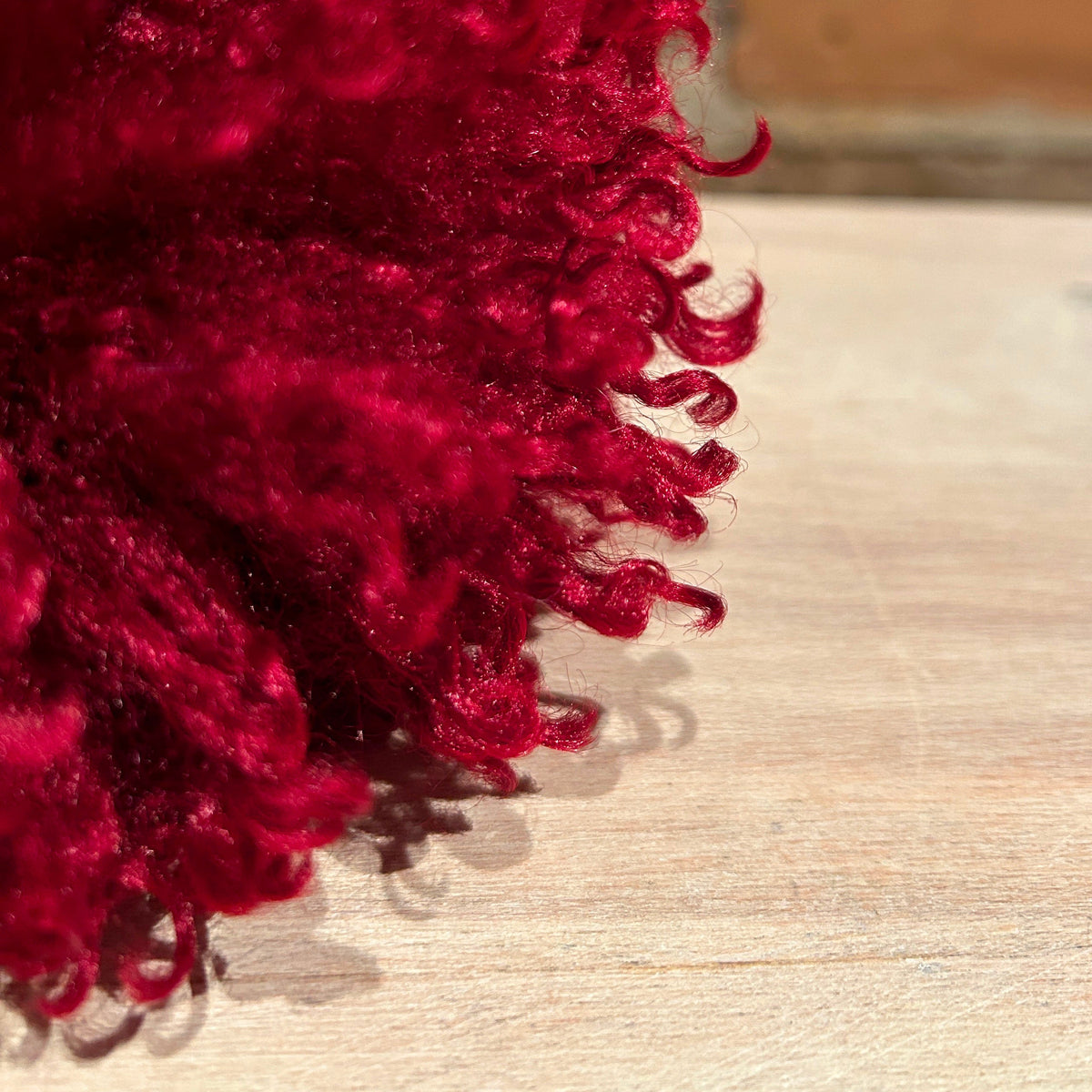 A close-up of a deep red curly pom-pom or fluffy decorative accessory resting on a light wood surface.