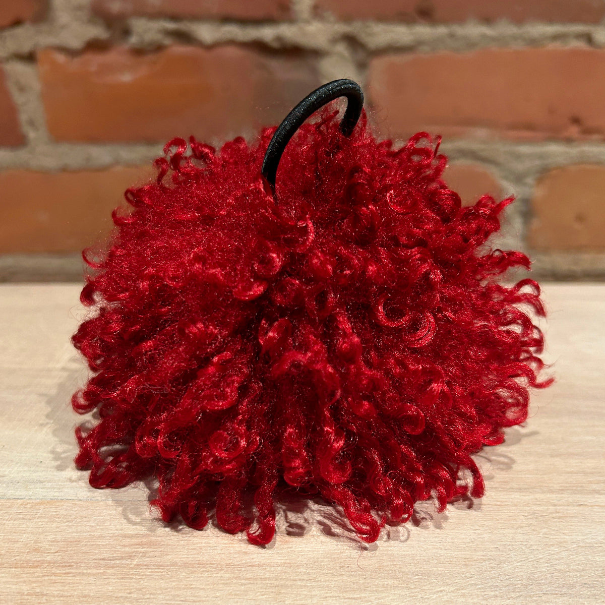 A bright red curly pom-pom accessory with a black elastic loop, resting on a light wood surface with a brick wall background.