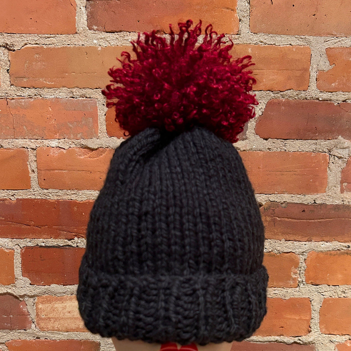 A dark charcoal gray knit beanie with a large, fluffy dark red pom-pom, displayed on a mannequin head against a red brick wall background.