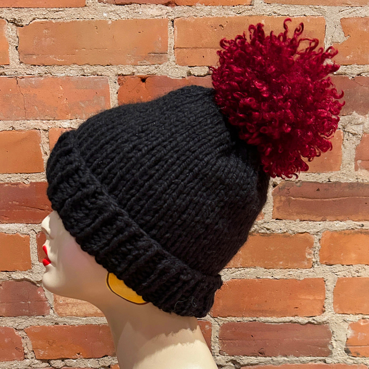 A black ribbed knit beanie with a large dark red curly pom-pom, displayed on a mannequin head against a red brick wall background.