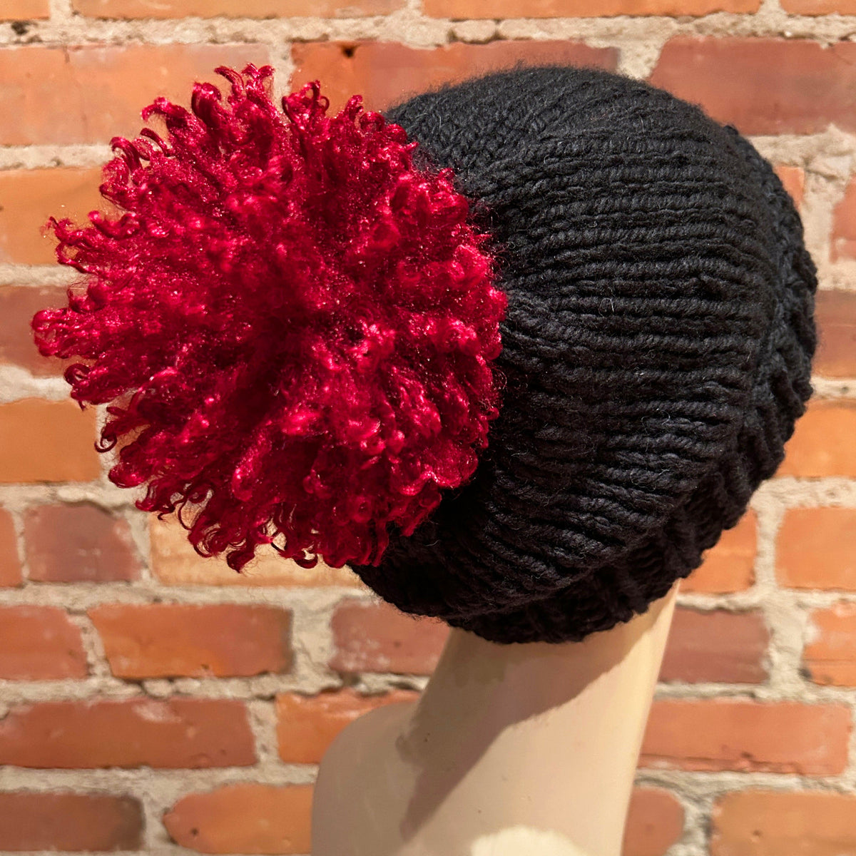 A black ribbed knit beanie hat with a large, fluffy red pom-pom, displayed on a mannequin head against a red brick wall background.