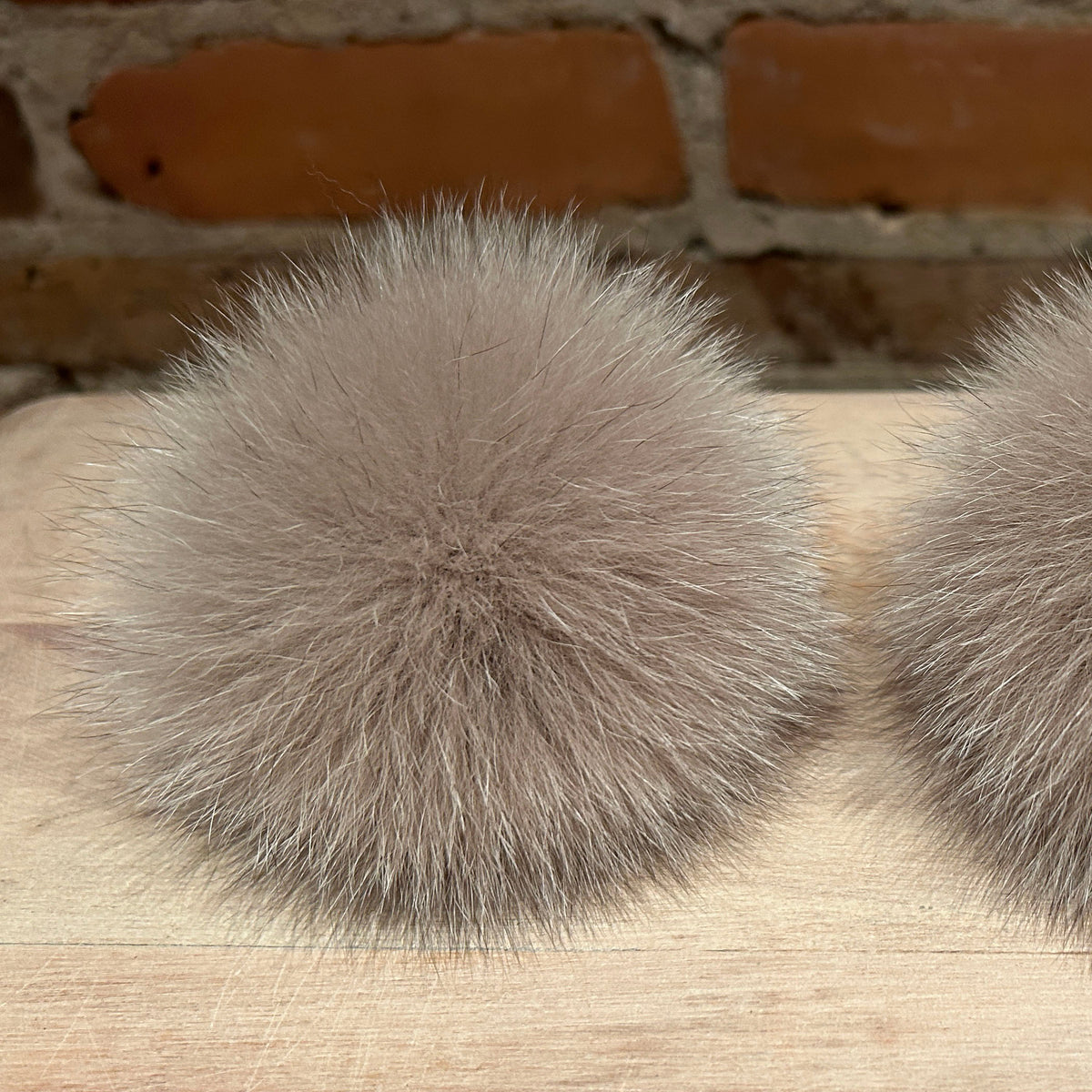 Two fluffy fox-fur pom-poms in a light taupe-gray color, resting on a light wood surface with a blurred brick wall background.
