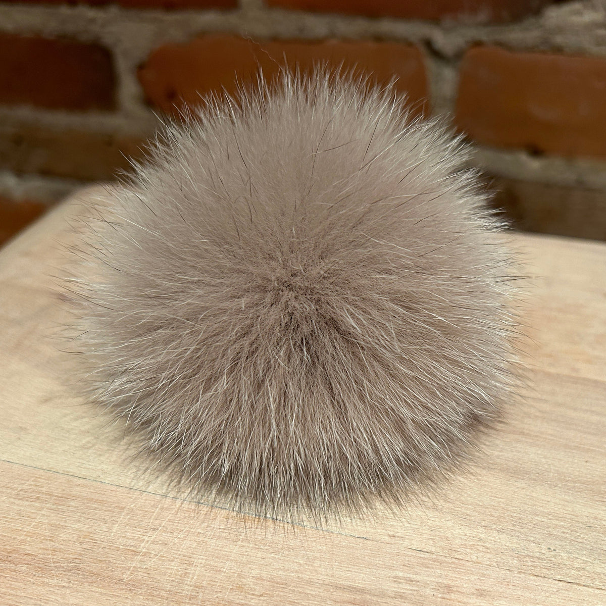 A fluffy pom-pom ball in a light taupe-gray color, resting on a light wood surface with a blurred brick wall background.