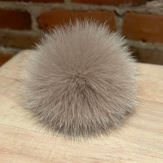 A fluffy pom-pom ball in a light taupe-gray color, resting on a light wood surface with a blurred brick wall background.