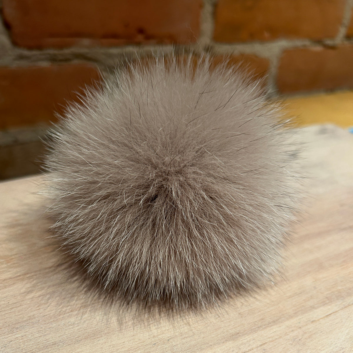 A fluffy pom-pom ball made of light taupe-gray fox fur, resting on a light wood surface with a brick wall background.