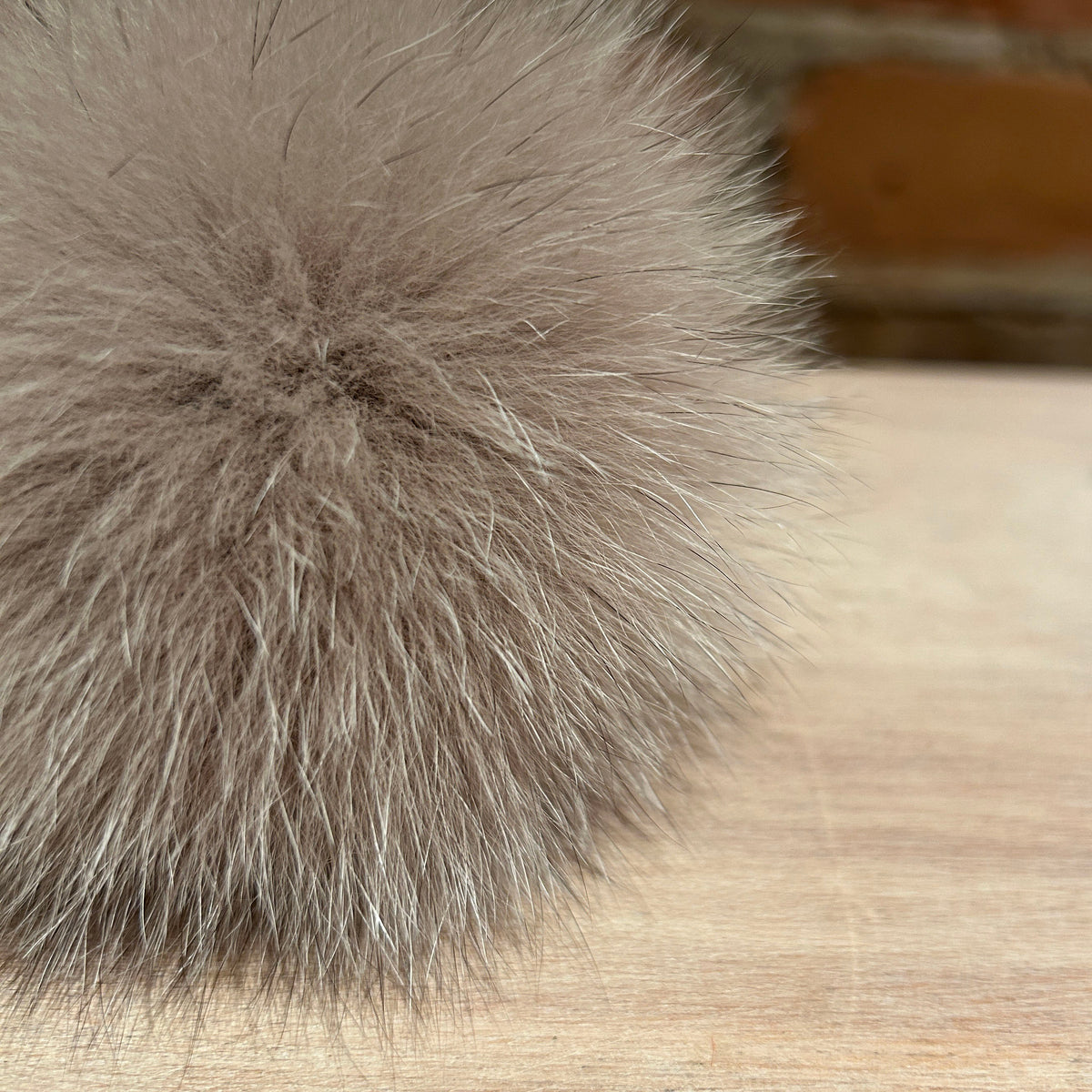 A close-up of a fluffy fox fur pom-pom in a taupe-gray color, resting on a light wood surface with a blurred brick wall background.