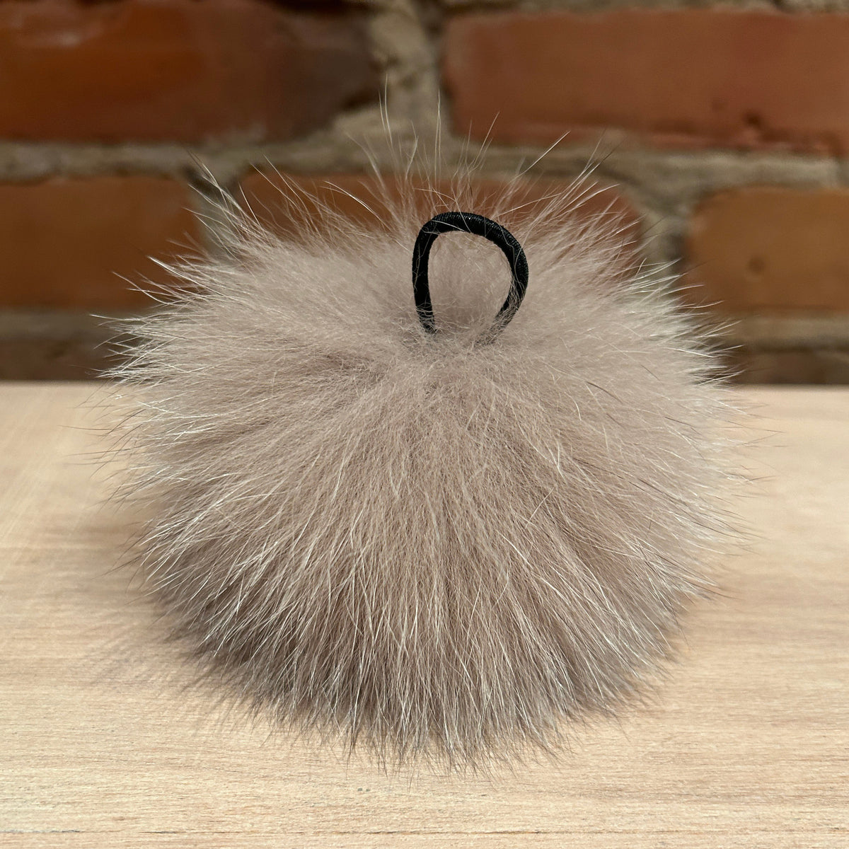 A fluffy fox-fur pom-pom keychain or bag charm in a light taupe-gray color, featuring a small black hanging loop at the top, resting on a light wood surface with a blurred brick wall background.