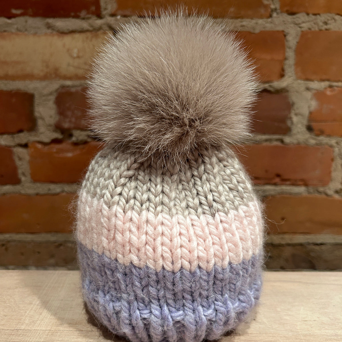 A hand-knit beanie hat with a large fluffy pom-pom, featuring horizontal stripes of gray, pale pink, lavender, and light blue, displayed on a wooden surface against a brick wall background.