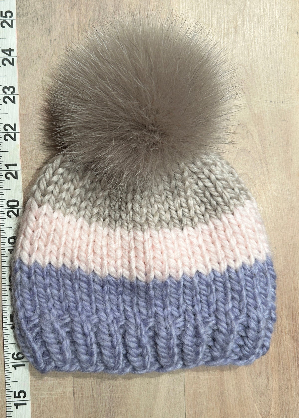 A small hand-knit beanie hat with horizontal stripes of taupe, pale pink, and dusty lavender, topped with a small fluffy taupe fox-fur pom-pom. A measuring tape is visible on the left side against a light wood background.