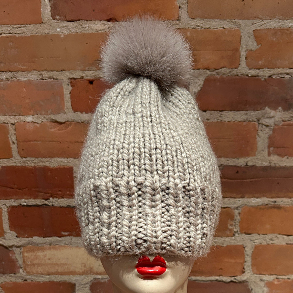 A light gray chunky knit beanie with a fluffy gray fox fur pom-pom, displayed on a mannequin head with red lips in front of a red brick wall.