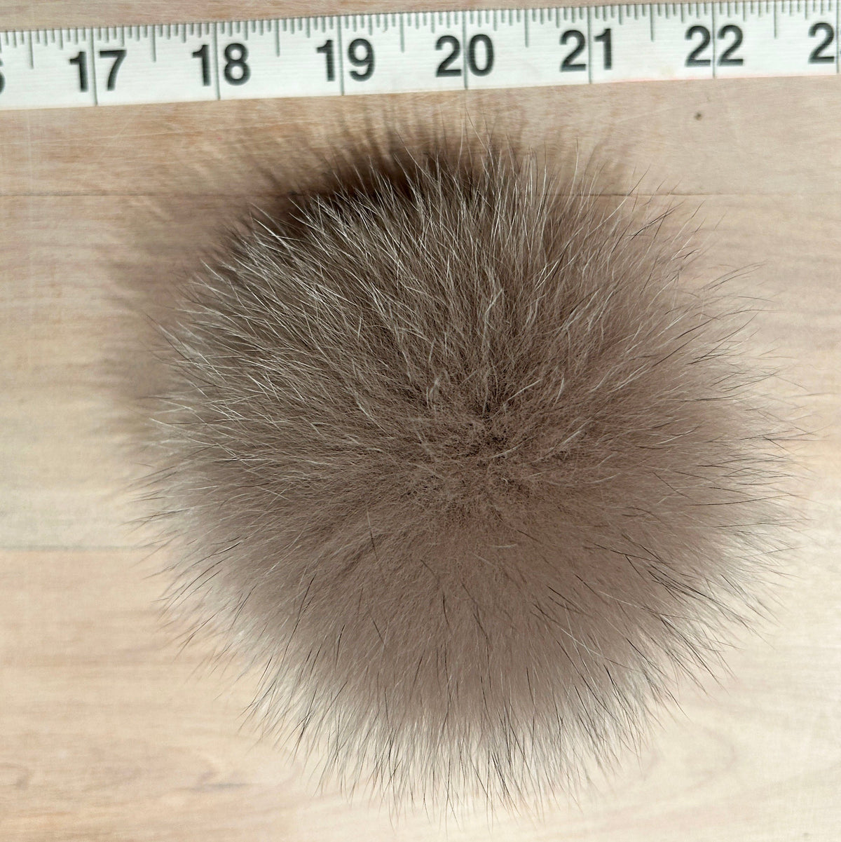 A round, fluffy fox fur pom-pom in a taupe-brown color, shown on a light wood surface with a measuring tape ruler at the top for scale.
