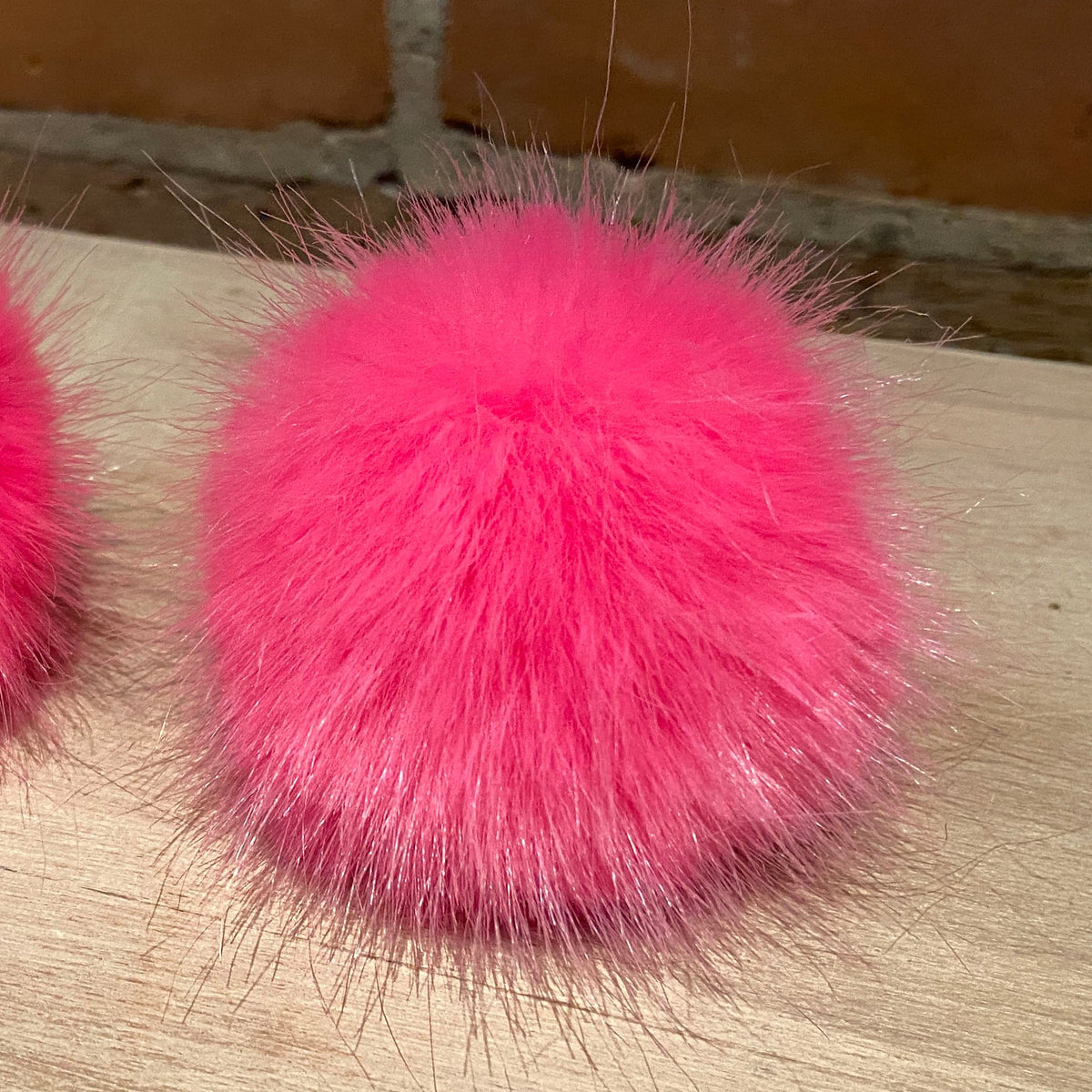 Two bright hot pink fluffy faux-fur pom-poms placed on a light wood surface with a brick wall background.