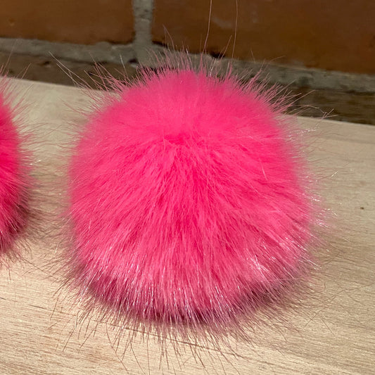 Two bright hot pink fluffy faux-fur pom-poms placed on a light wood surface with a brick wall background.