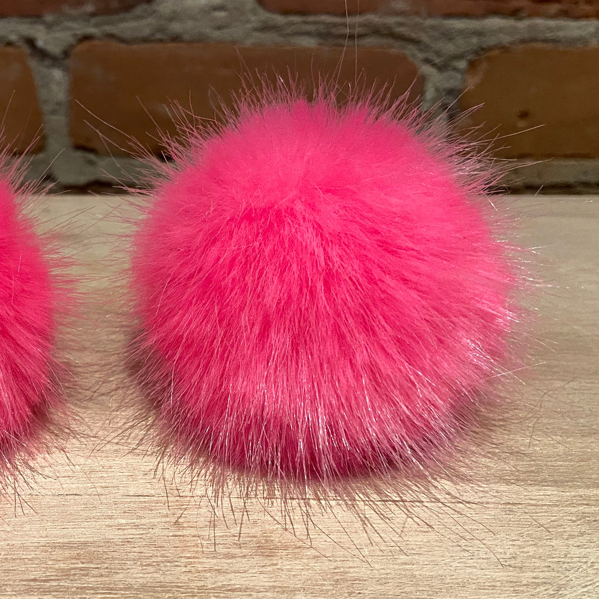Two bright pink fluffy faux-fur pom-poms placed side by side on a light wood surface with a brick wall background.