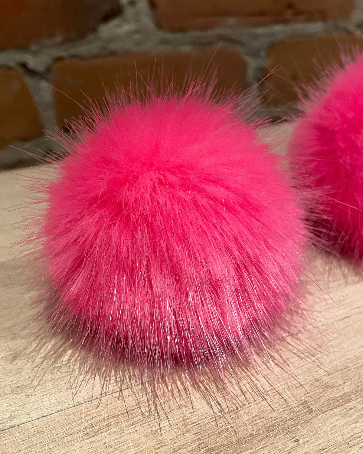 Two bright pink fluffy faux-fur pom-poms resting on a light wood surface with a blurred brick wall in the background.