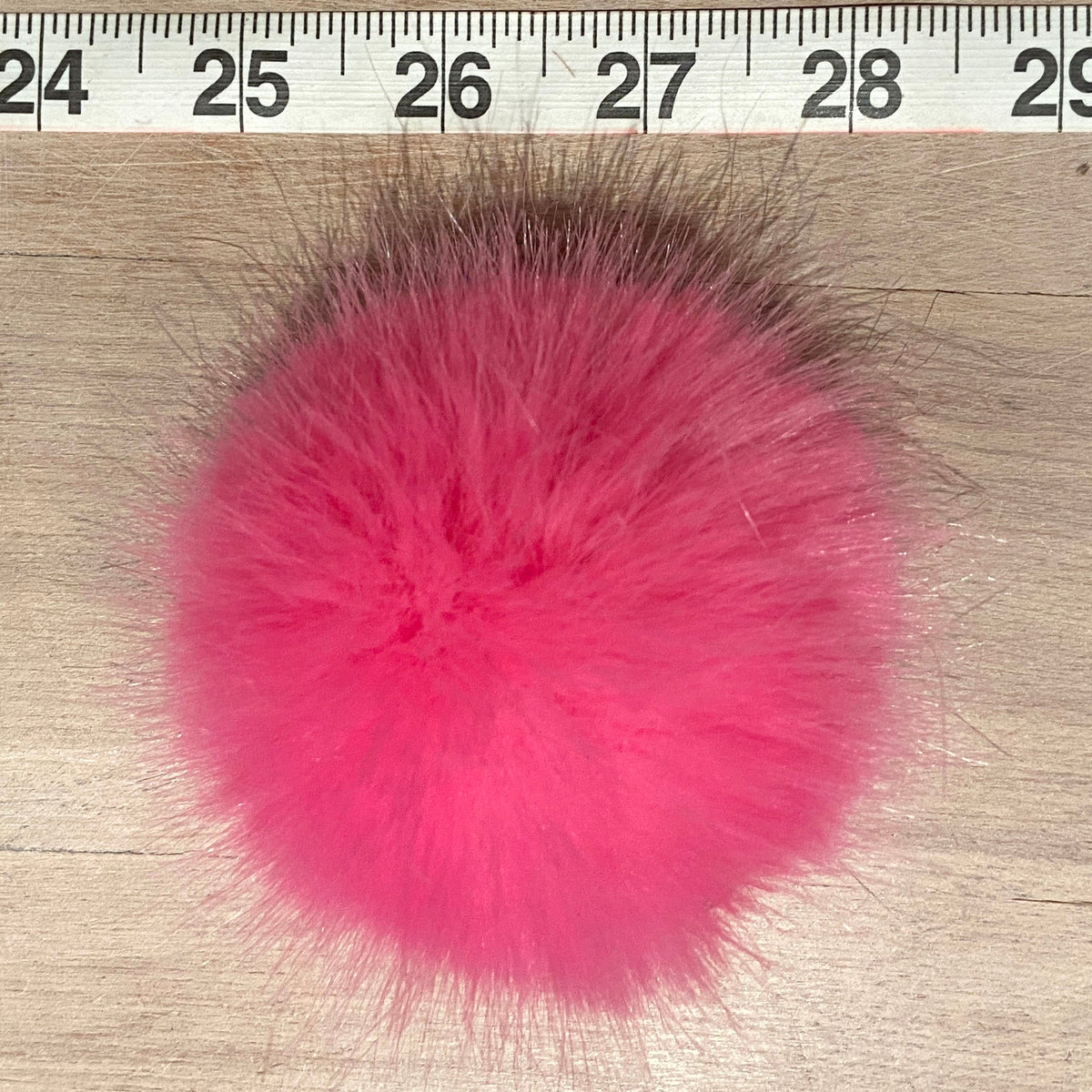A bright pink fluffy faux fur pom-pom shown on a light wood surface with a measuring ruler at the top for scale.