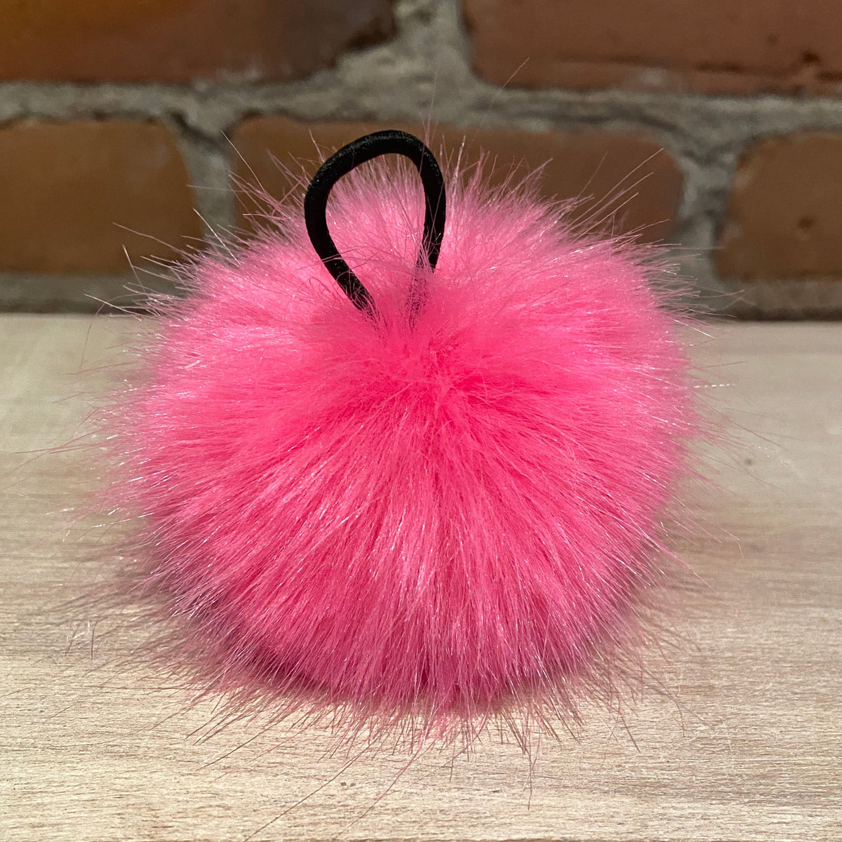A bright pink fluffy pom-pom with a black elastic loop, resting on a light wood surface with a brick wall background.